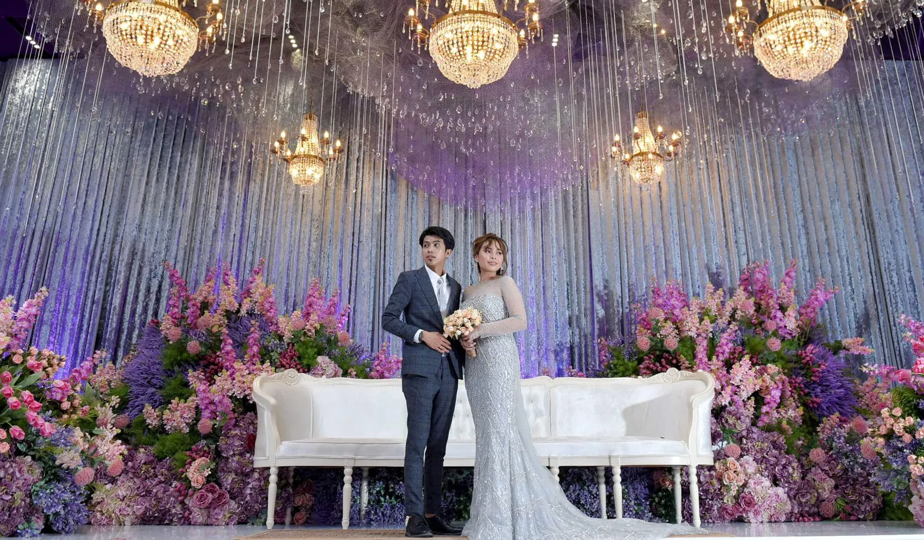 Pasangan pengantin di pentas dengan candelier kristal dan bunga ungu