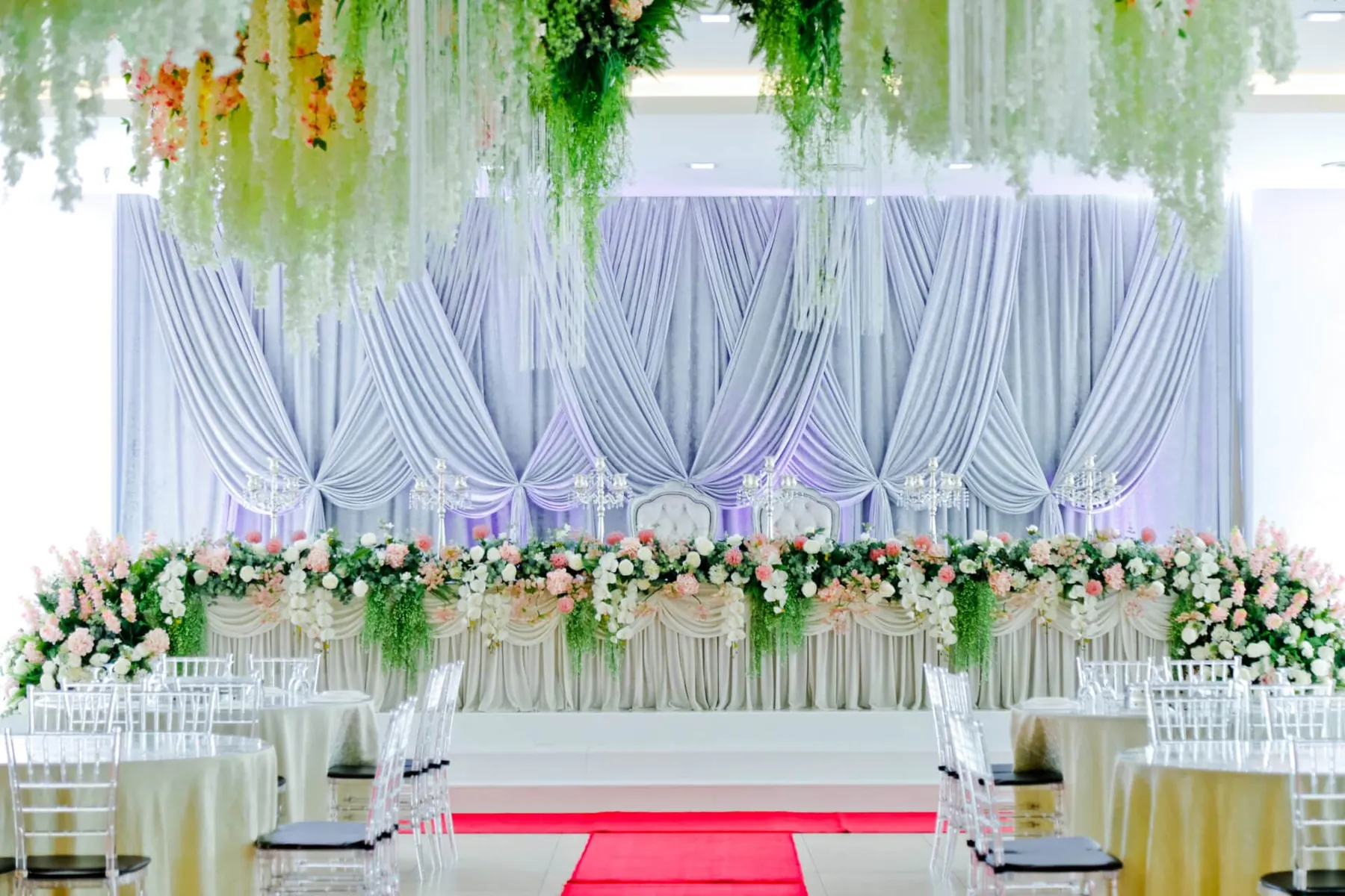 Pelamin putih berhias bunga hijau dengan meja tetamu dan laluan permaidani merah